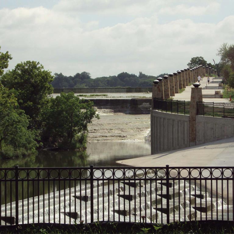 View of a White Rock Lake's dam spillway with water flowing over stepped concrete structures, surrounded by trees and a pathway with lamp posts.