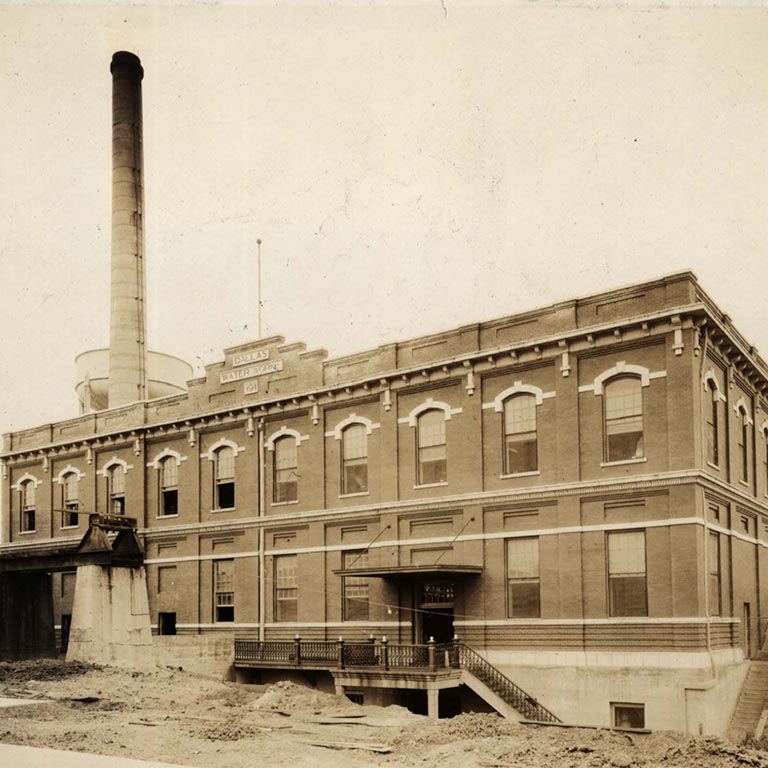 Sepia-toned historic photo of a large brick industrial building labeled 'Dallas Water Works' with a tall smokestack at White Rock Lake.