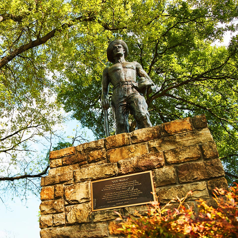 Bronze statue of a shirtless CCC worker holding a tool and hat atop a stone pedestal with a plaque, surrounded by green trees at White Rock Lake.