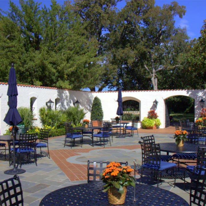 Outdoor patio with black metal tables and chairs topped with orange flower pots, surrounded by white stucco walls and greenery under a clear blue sky.