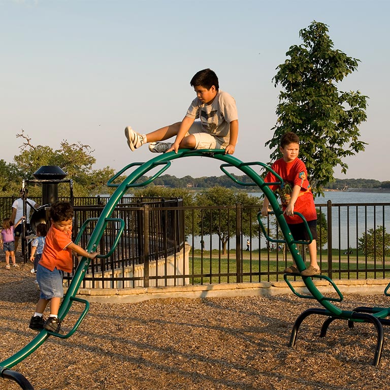 Three children playing on a green climbing arch at a park playground near a lake in the late afternoon.