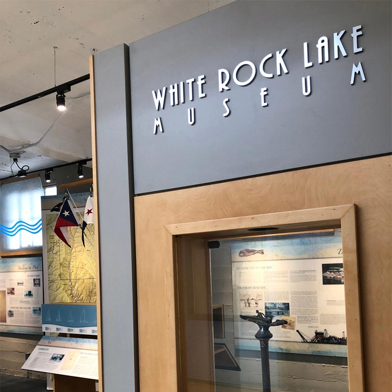 Exterior of White Rock Lake Museum featuring exhibits, a map, and Texas flags on display.