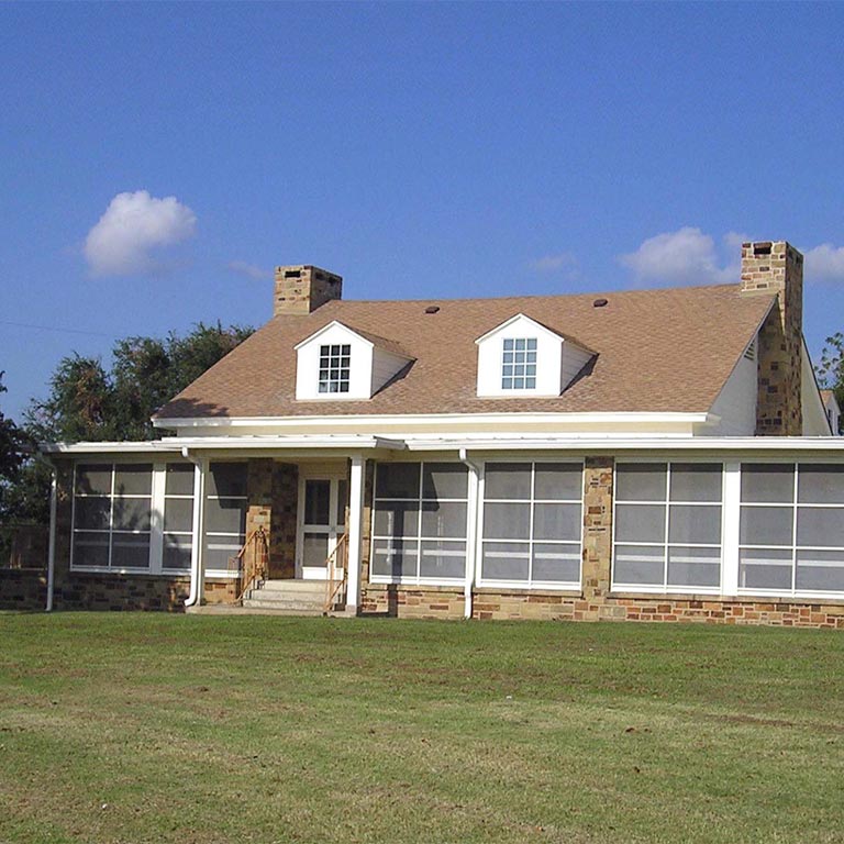 Single-story house with stone chimney pillars, screened-in porch, and two dormer windows under a clear blue sky.