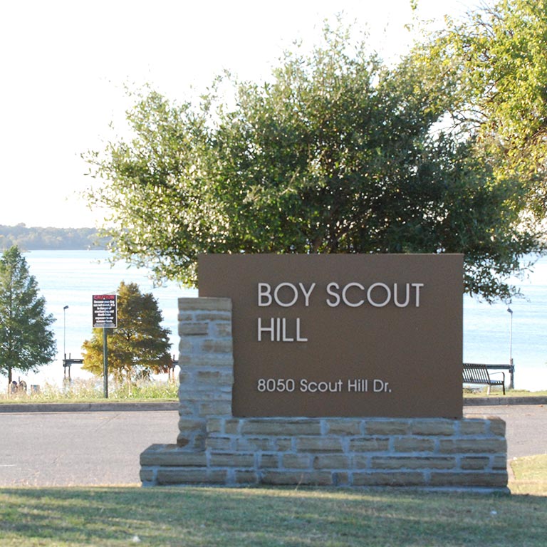 Brown stone sign reading 'Boy Scout Hill, 8050 Scout Hill Dr.' with trees and a lake in the background.