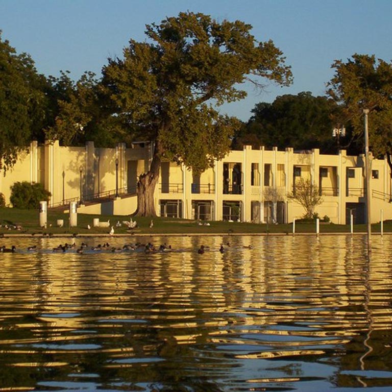 White Rock Lake's bath house with trees and birds near the water's edge, reflecting in the rippling water.