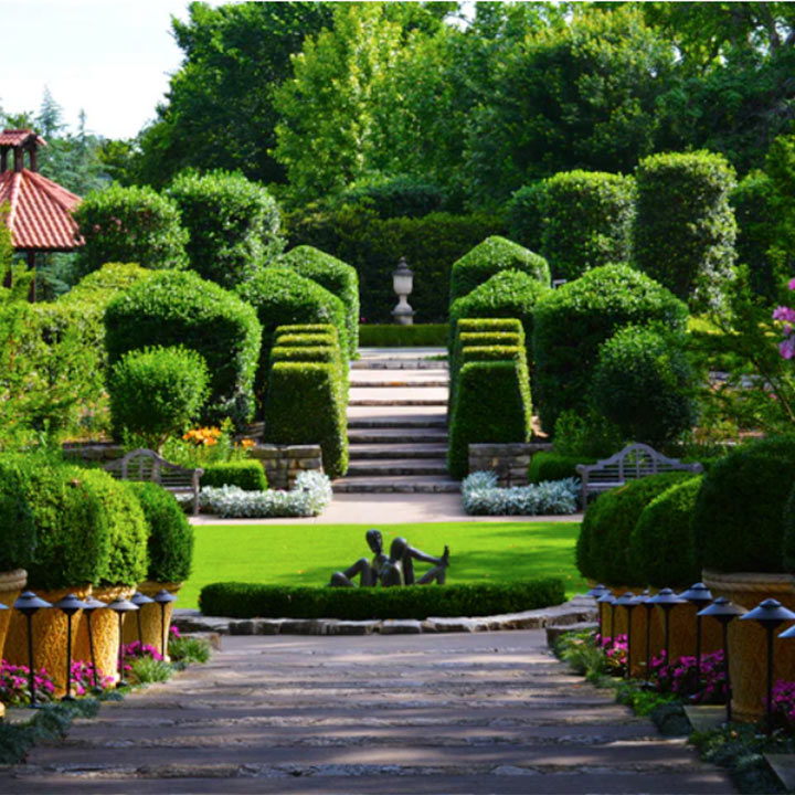 Pathway leading through manicured garden with trimmed hedges, stone steps, and a bronze sculpture of two reclining figures on grass.