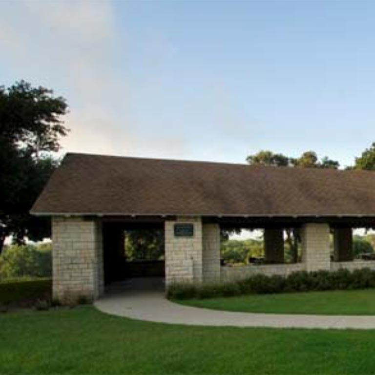Stone pavilion with a brown shingled roof and open sides next to a curved sidewalk on a grassy hill under a blue sky.