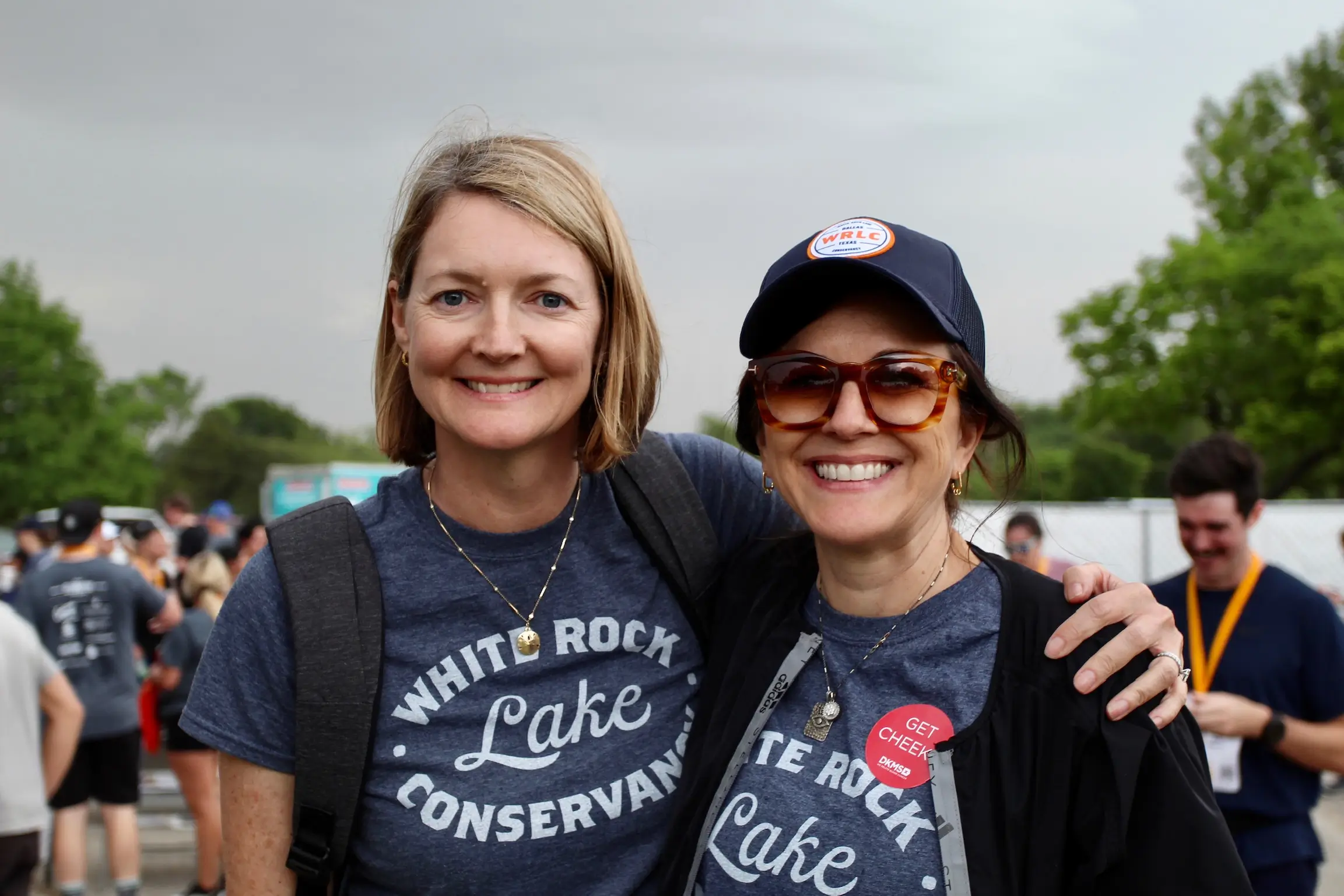Two smiling women wearing matching White Rock Lake Conservancy t-shirts at an outdoor event.