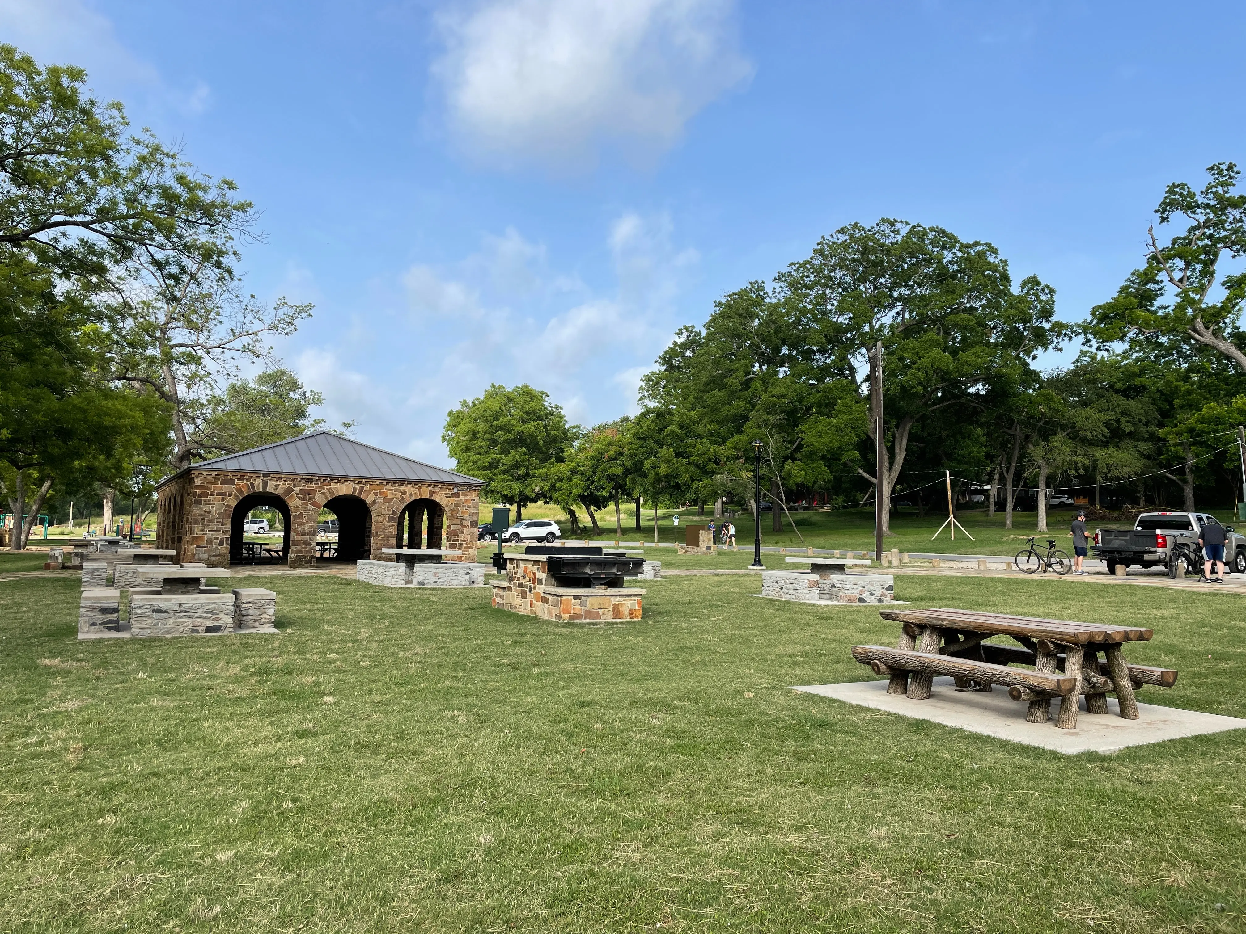 Park scene with stone picnic tables, a stone pavilion with arches, green grass, and trees at White Rock Lake Park in Dallas.