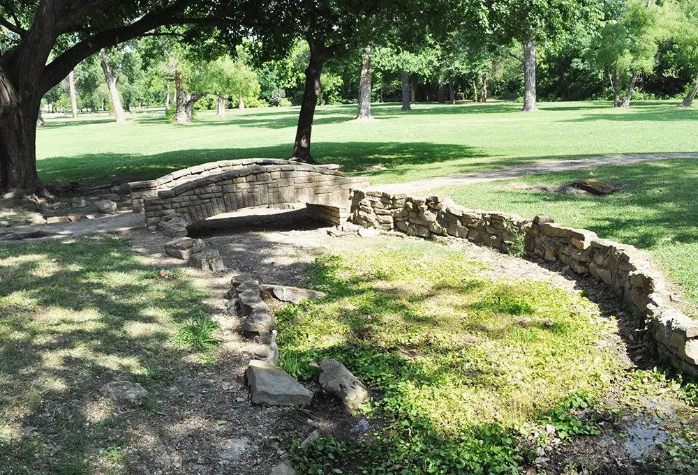 Small stone footbridge over a shallow, dry creek bed surrounded by green grass and trees at White Rock Lake Park in Dallas.