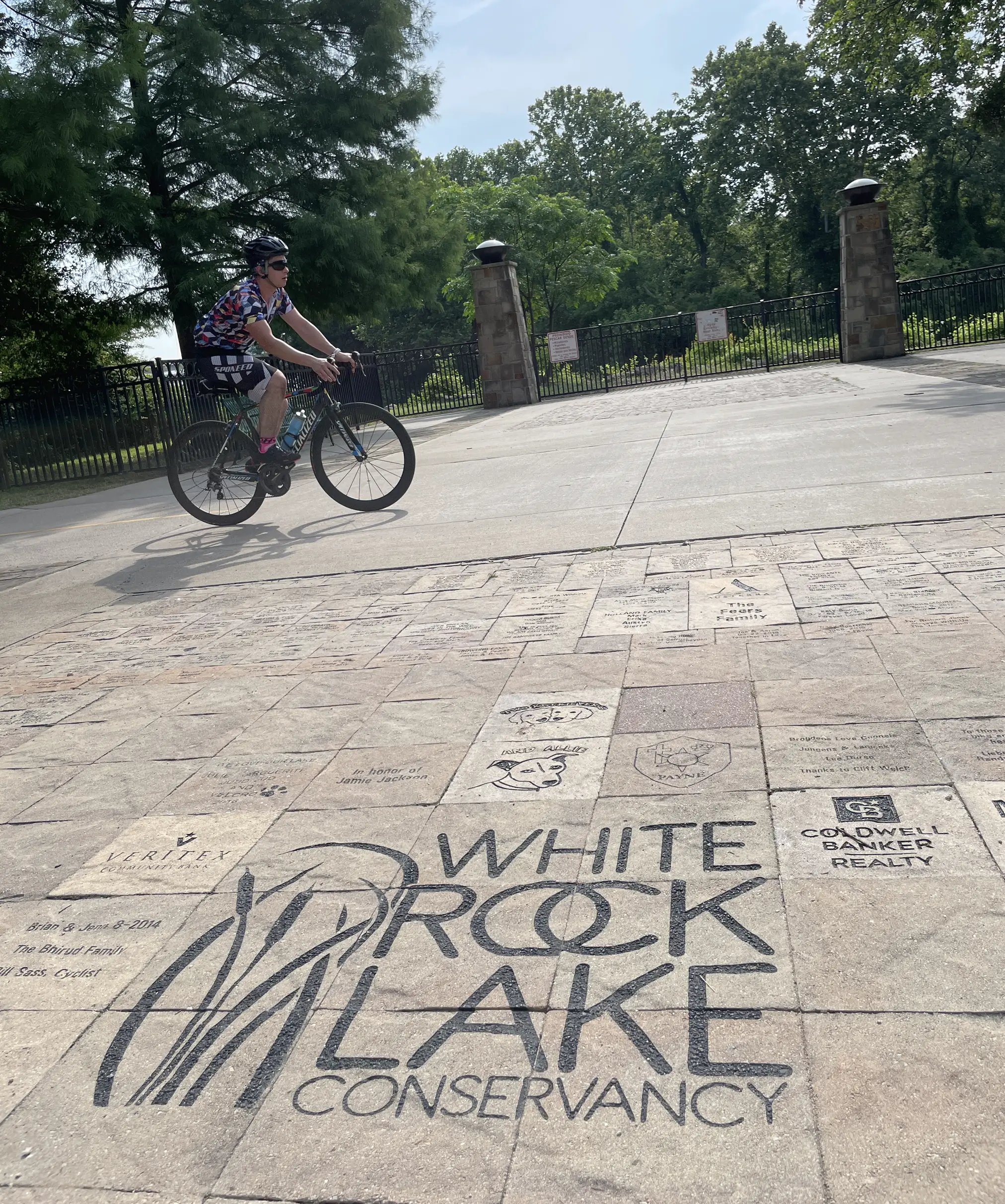 Cyclist wearing a helmet riding past a paved area with White Rock Lake Conservancy logo and engraved bricks, surrounded by trees and stone pillars.
