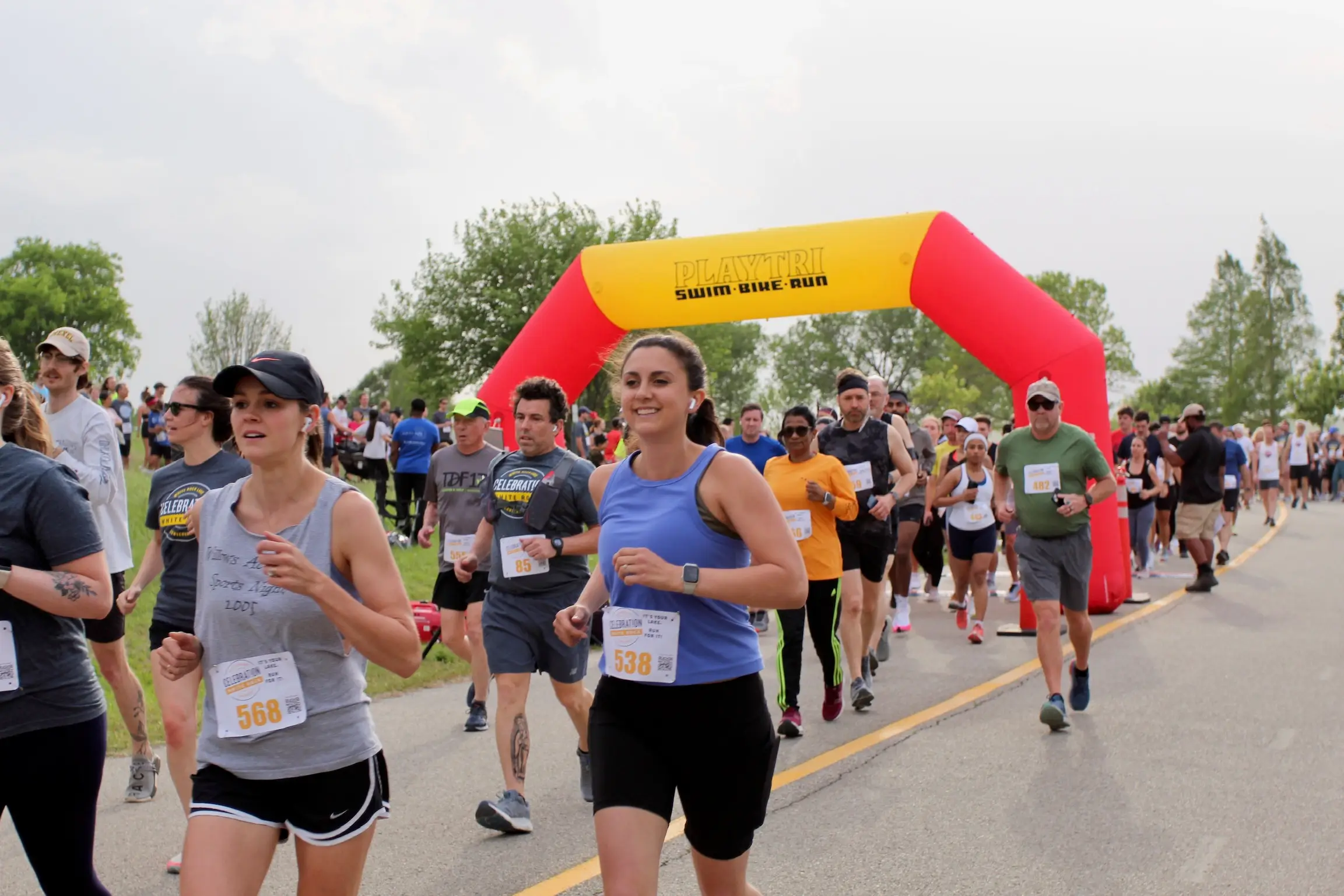 Celebration White Rock Dallas Runners participating in an outdoor race passing under a red and yellow inflatable arch labeled 'PLAYTRI Swim Bike Run