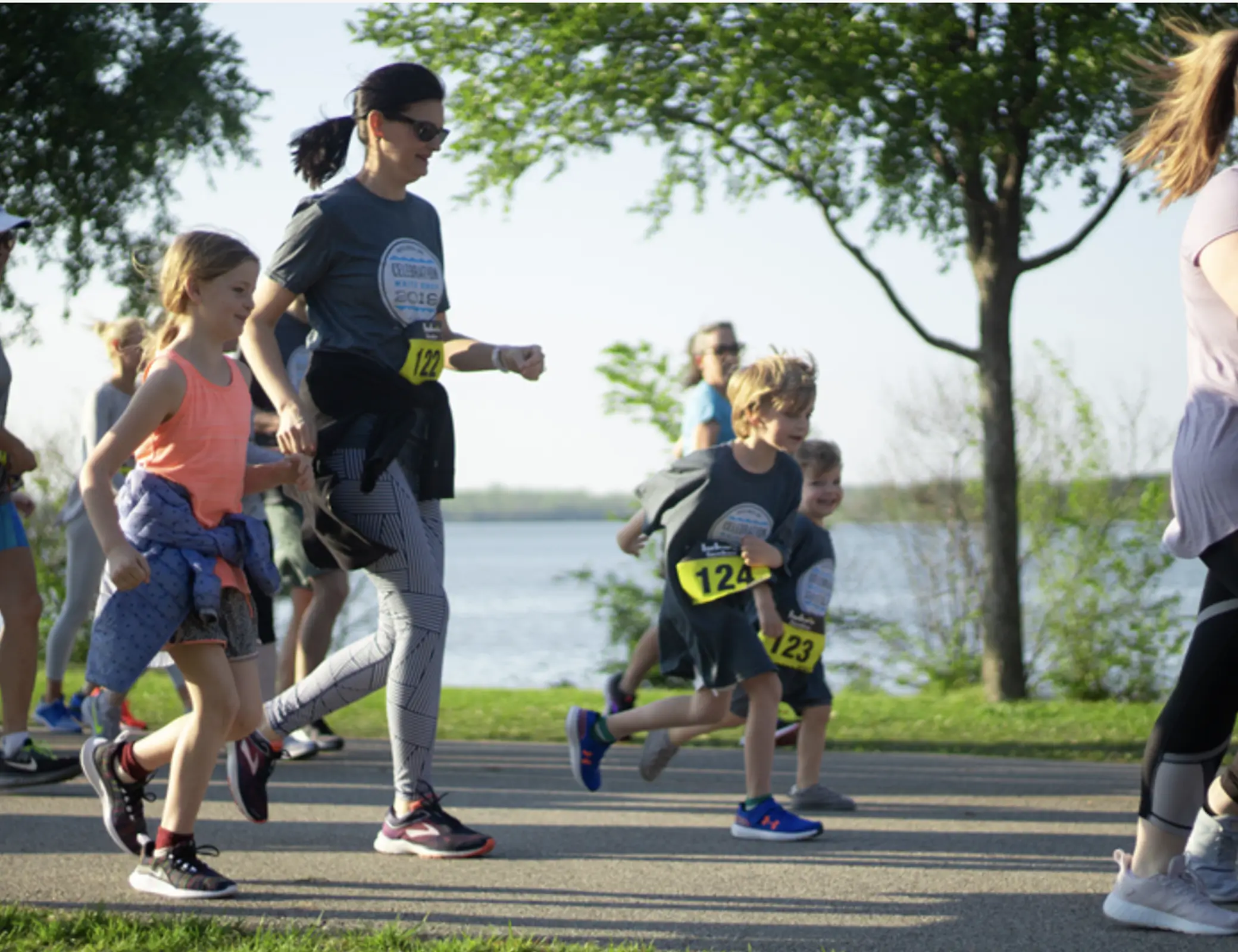 Group of children and adults running in a race near a lake with trees and grass in the background on a sunny day at Celebration White Rock race in Dallas, TX.