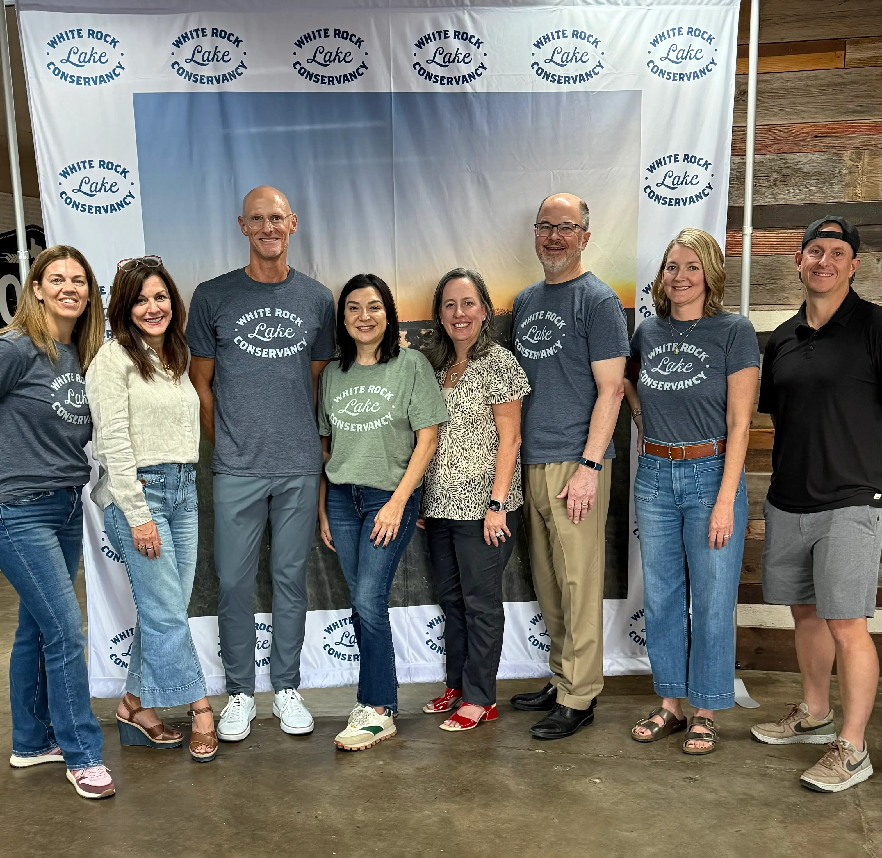 Eight people standing indoors smiling in front of a White Rock Lake Conservancy backdrop.