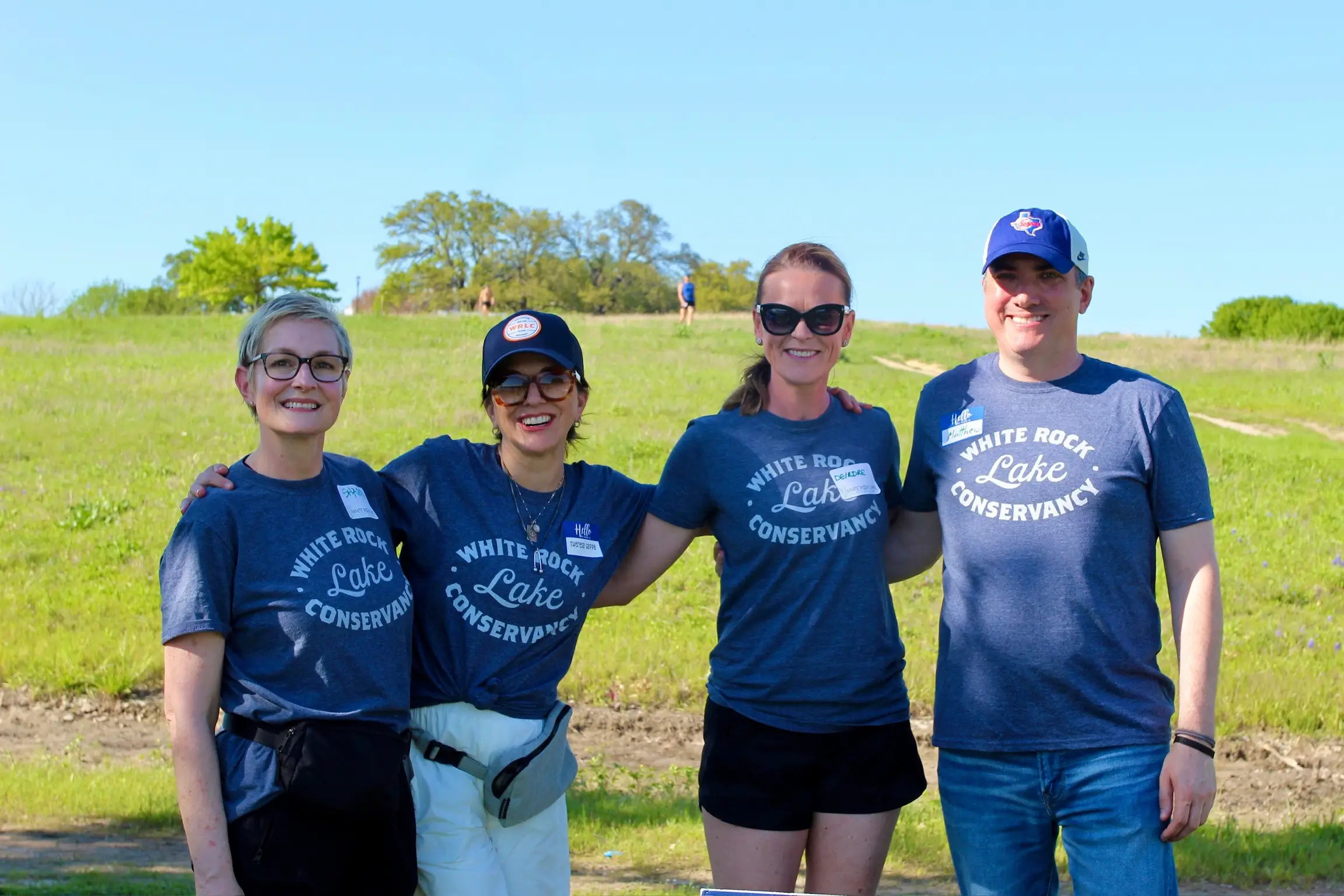 Four smiling adults wearing matching Blue 'White Rock Lake Conservancy' T-shirts stand with arms around each other in a sunny grassy field.