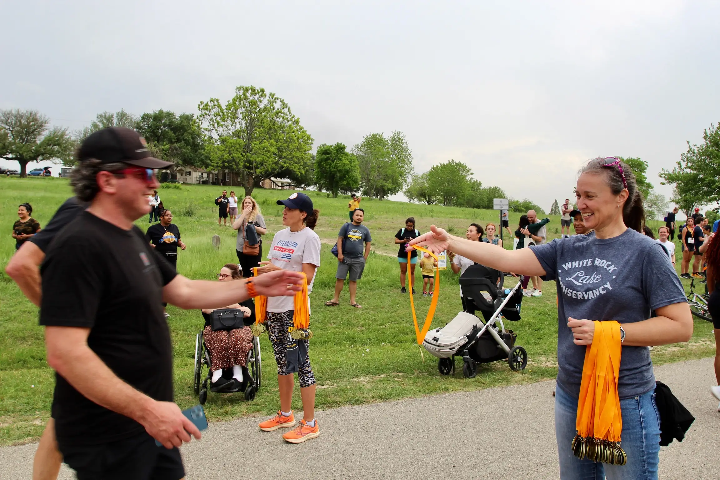 Smiling woman wearing a White Rock Lake Conservancy t-shirt handing yellow race medals to participants at an outdoor event with people milling around on grass.