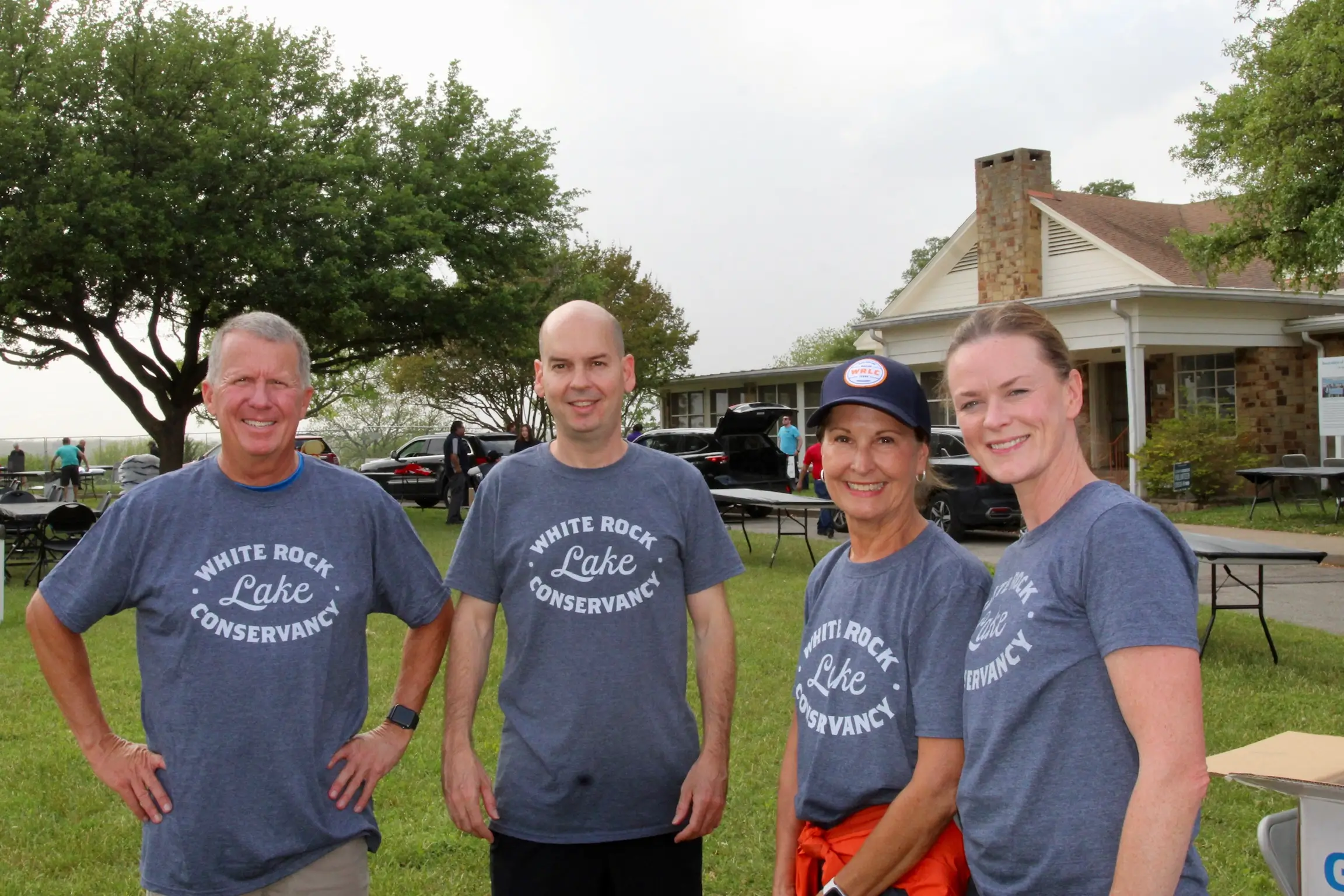 Four adults wearing matching Gray White Rock Lake Conservancy t-shirts standing outside on grass with trees and a building in the background.