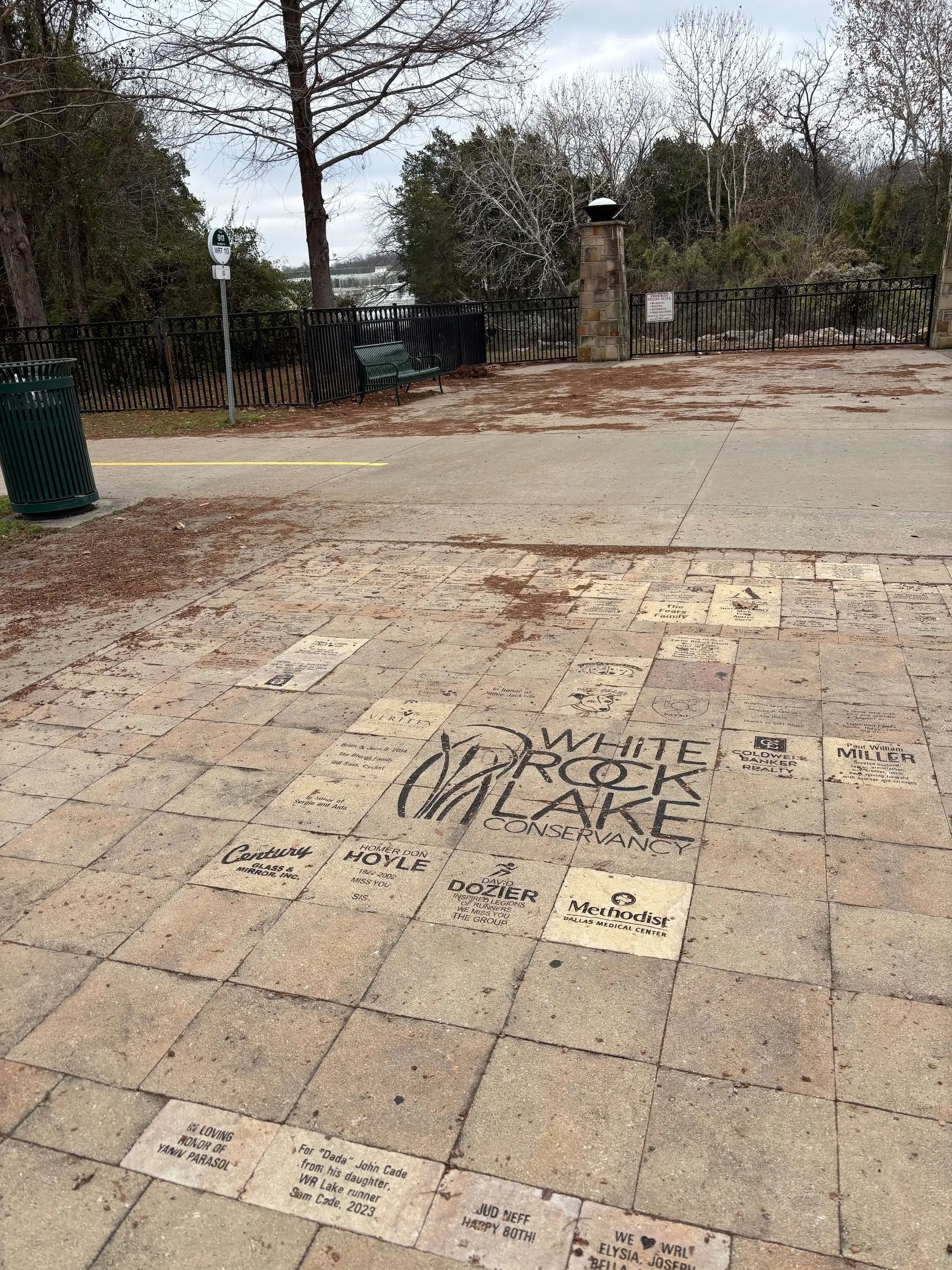 Paved area with engraved commemorative bricks including a large White Rock Lake Conservancy logo, surrounded by trees and a fenced area with a bench.