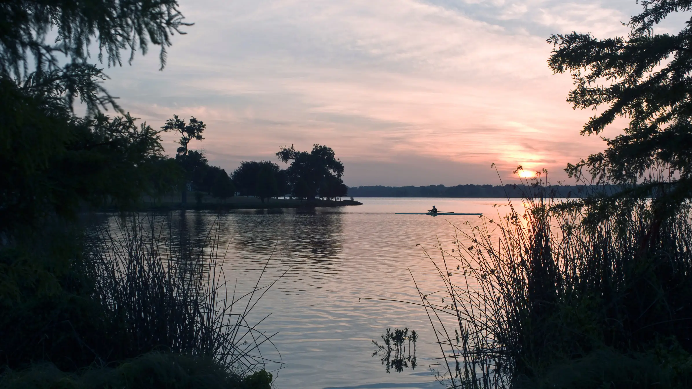 Person rowing a kayak on a calm lake at sunset with silhouettes of trees and reeds framing the water.