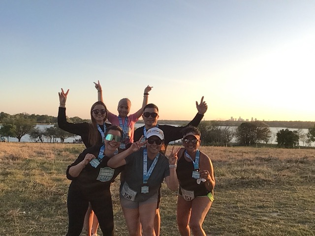 Group of six runners smiling and posing with race medals near a lake during sunset.