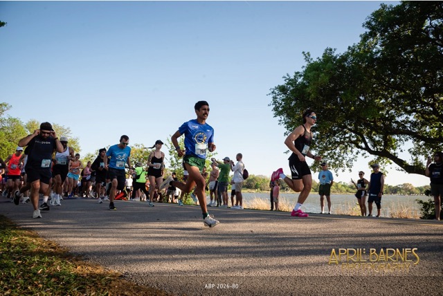 Runners participating in a race outdoors on a sunny day with spectators by a tree and a body of water.