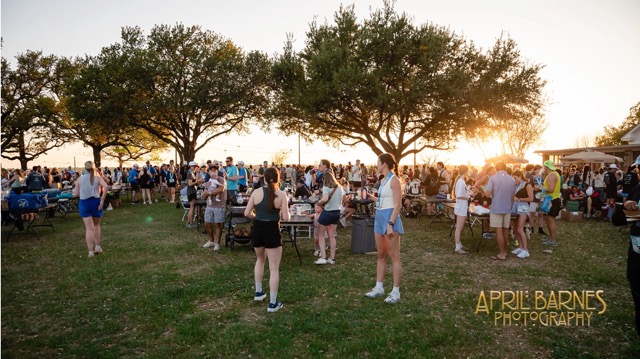 Large group of people gathered outdoors on grass near trees during sunset, some standing and some seated at tables, likely at a race event.