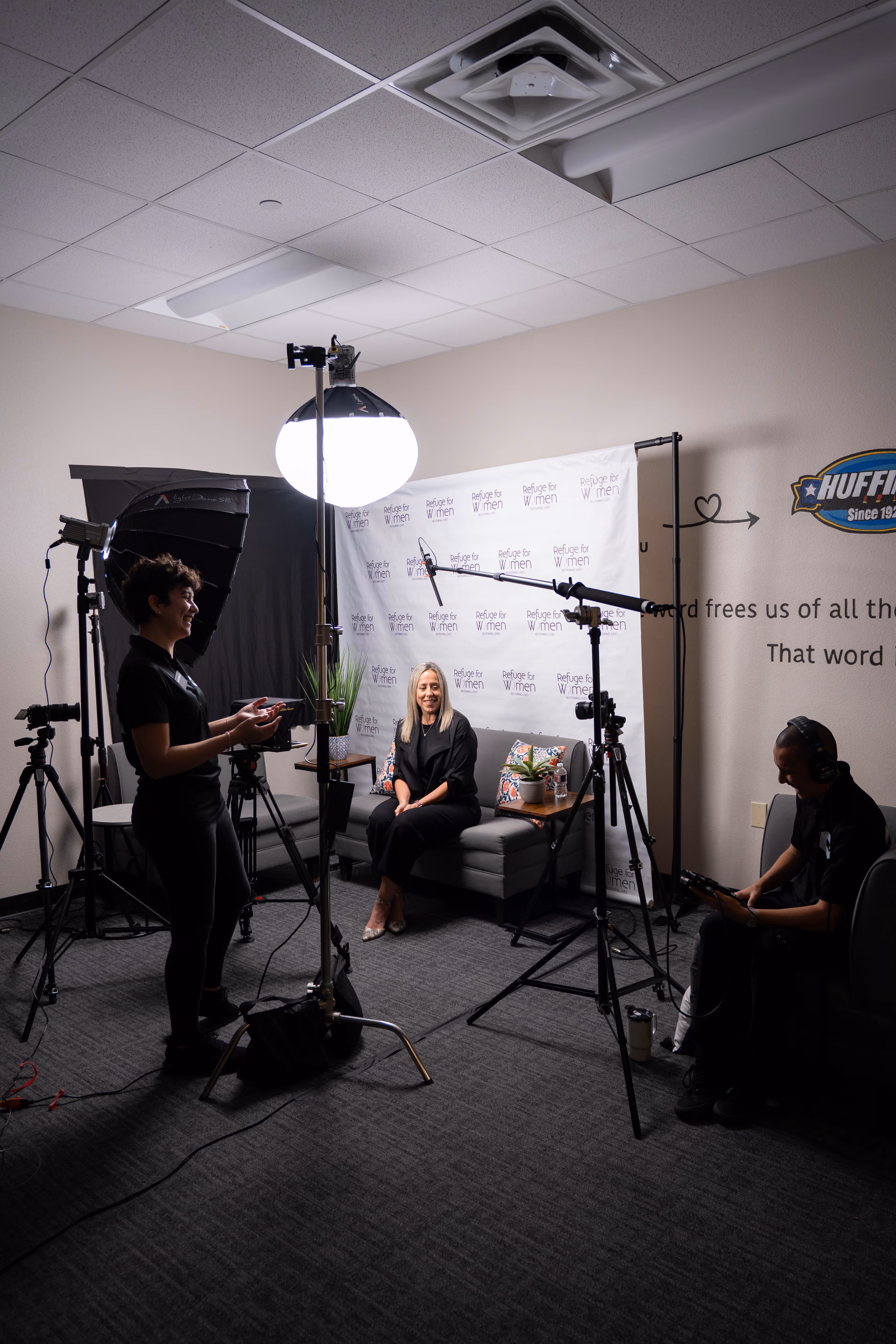 A woman sits on a gray couch smiling for an interview with cameras, lighting equipment, and two crew members in a small studio room.