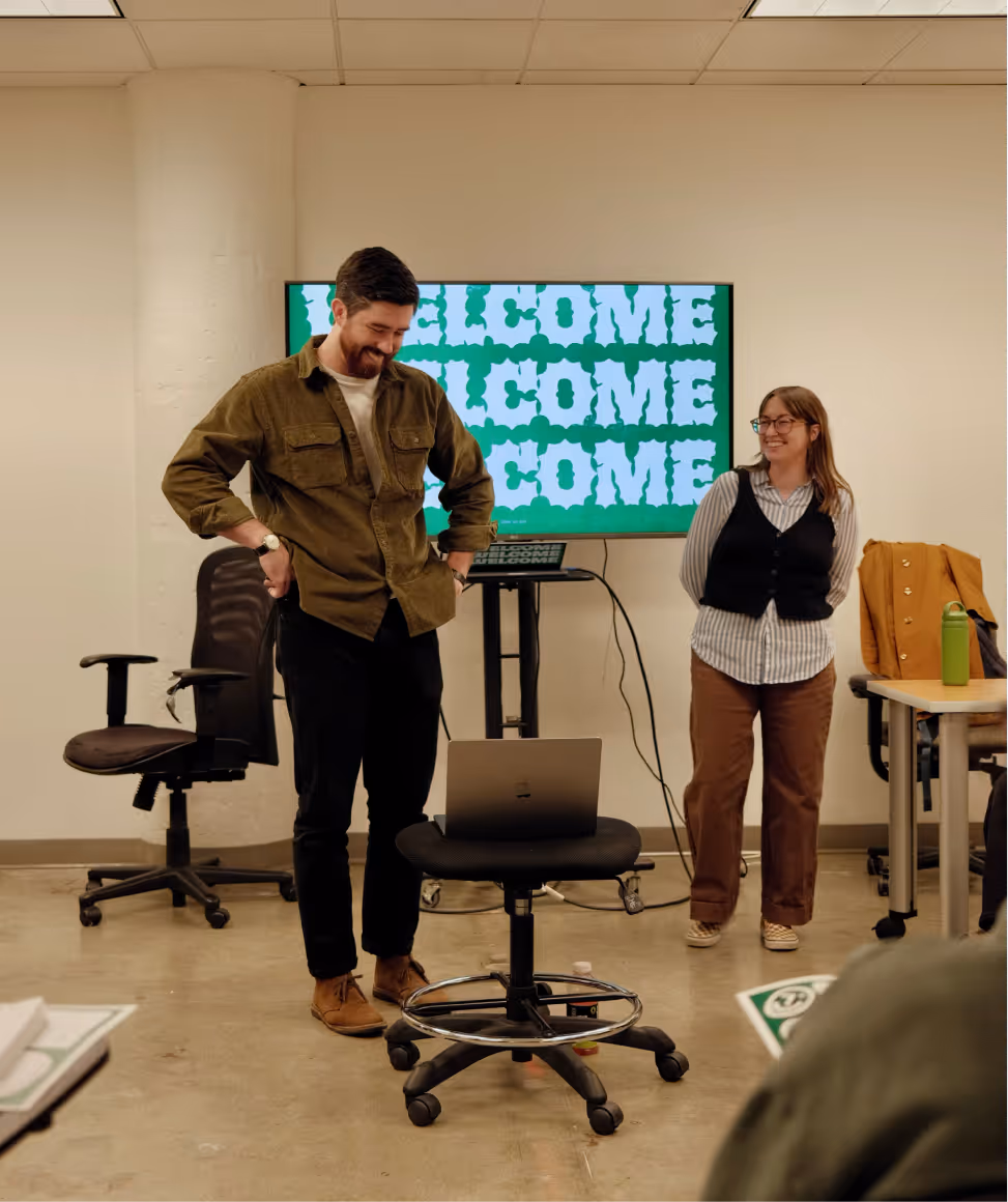 Man and woman standing in a room with a laptop on a chair and a screen behind displaying the word 'WELCOME' repeatedly.