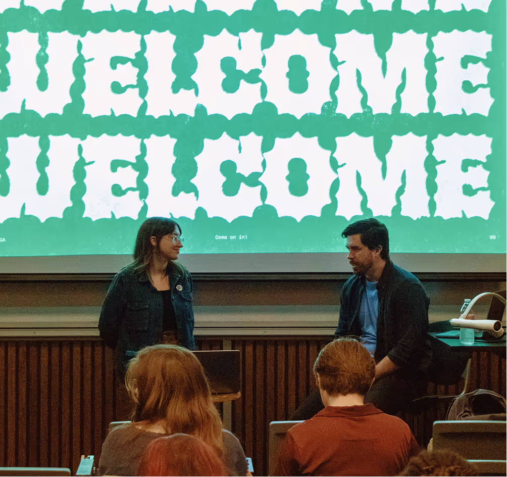 Two people, a woman and a man, engaged in conversation in front of a large green screen displaying the word 'WELCOME' twice, with an audience seated watching them.