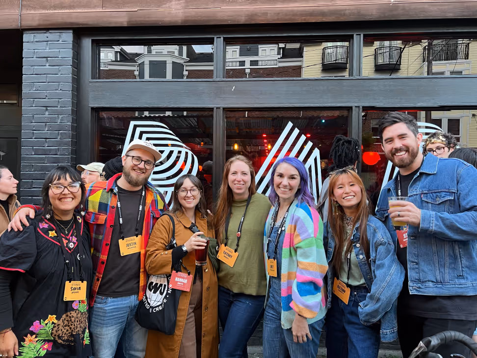 Group of seven smiling young adults standing close together outside a building with large windows, some holding drinks.