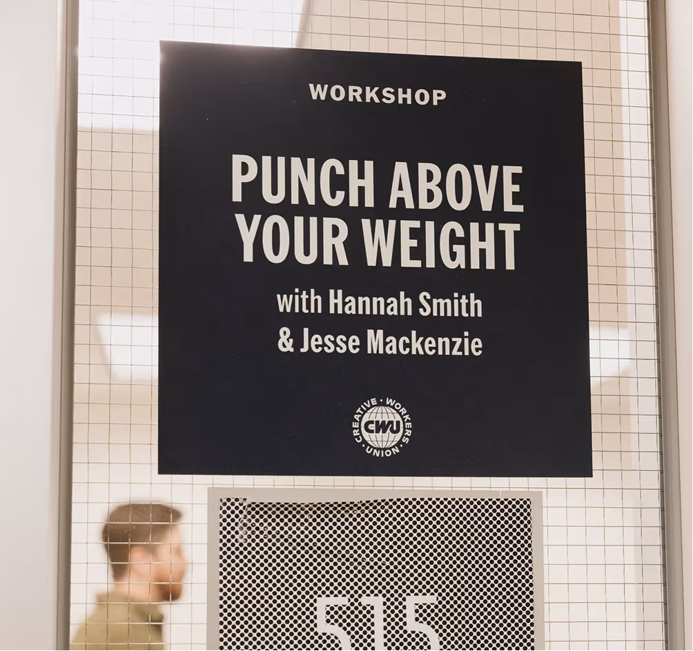 Black poster on a window with text: Workshop Punch Above Your Weight with Hannah Smith & Jesse Mackenzie, and a blurred man walking behind it.