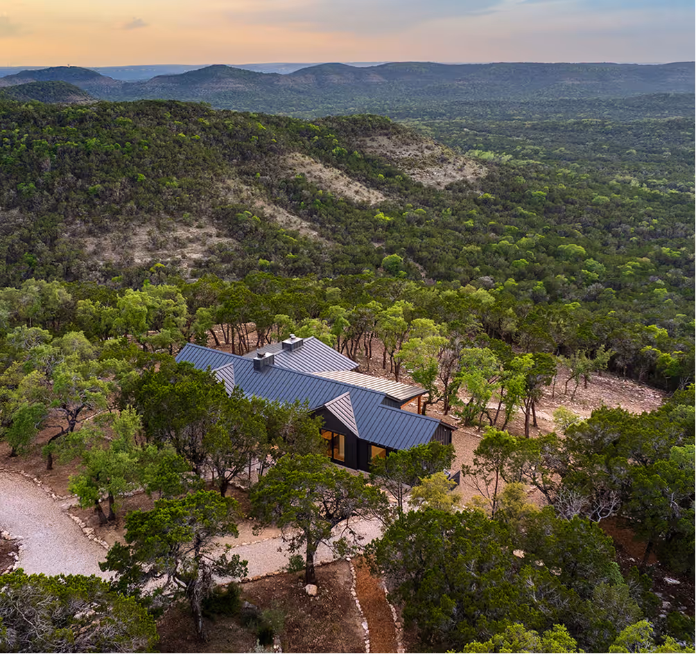 Modern house with a dark roof surrounded by dense green trees in a hilly forested landscape at sunset.