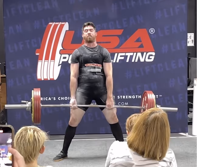 Jesse in a black singlet lifting a 465lb barbell on a stage with USA Powerlifting banner in the background.