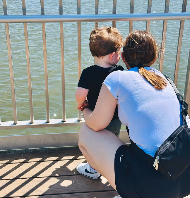 Woman crouching and holding a toddler as they look out over a body of water through metal railings.