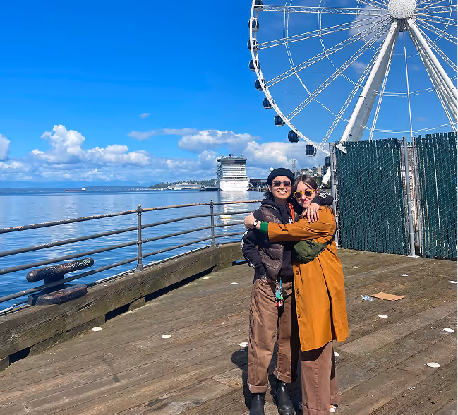 Two women hugging on a wooden pier with a large Ferris wheel, ocean, and cruise ship in the background under a blue sky.