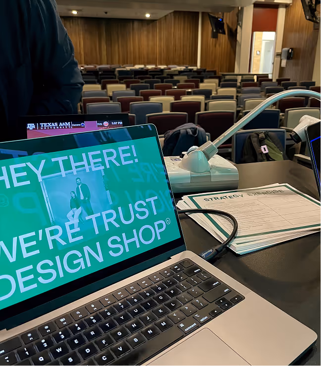 Laptop displaying text 'HEY THERE! WE'RE TRUST DESIGN SHOP' on a desk in an empty lecture hall with multicolored chairs.