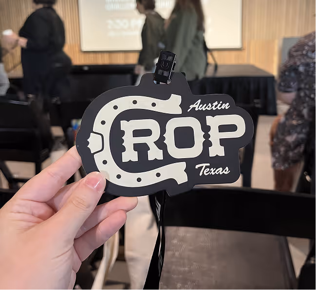 Hand holding a black badge for Chop Con in Austin, featuring a horseshoe design, in a room with people and chairs.