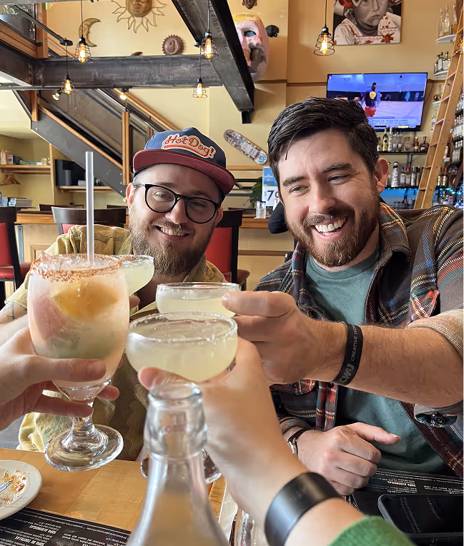 Jesse and Justin smiling and clinking cocktails with others in a lively bar interior featuring warm lighting and wall decorations.