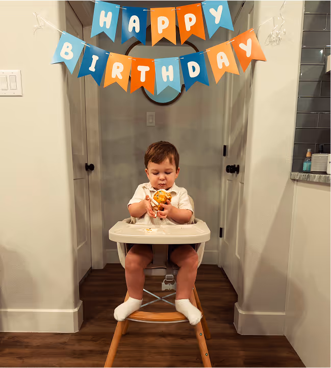 Toddler sitting in a high chair holding a partially eaten cupcake under a blue and orange Happy Birthday banner.