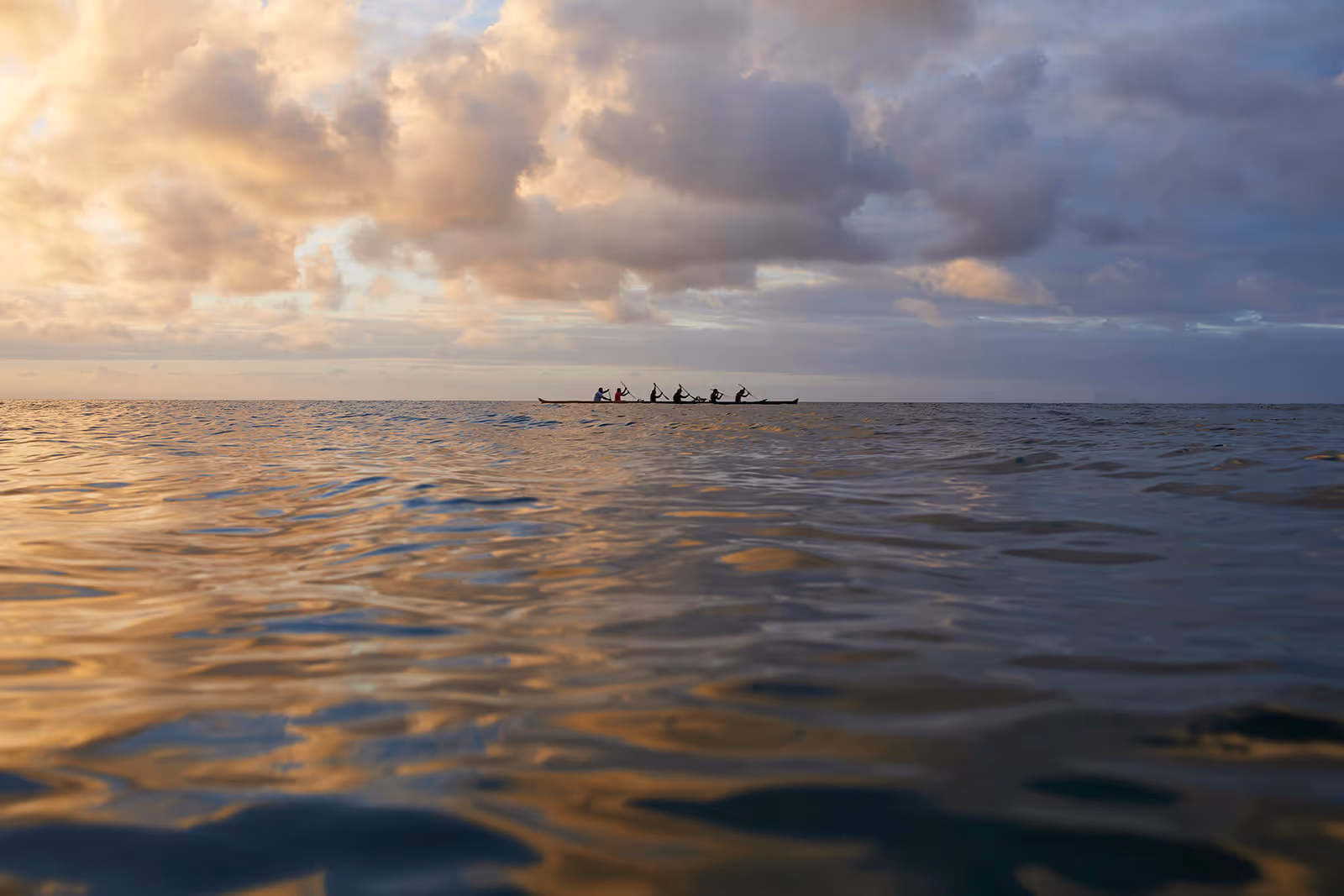 Six people paddling a long canoe on calm water under a cloudy sky during sunset.