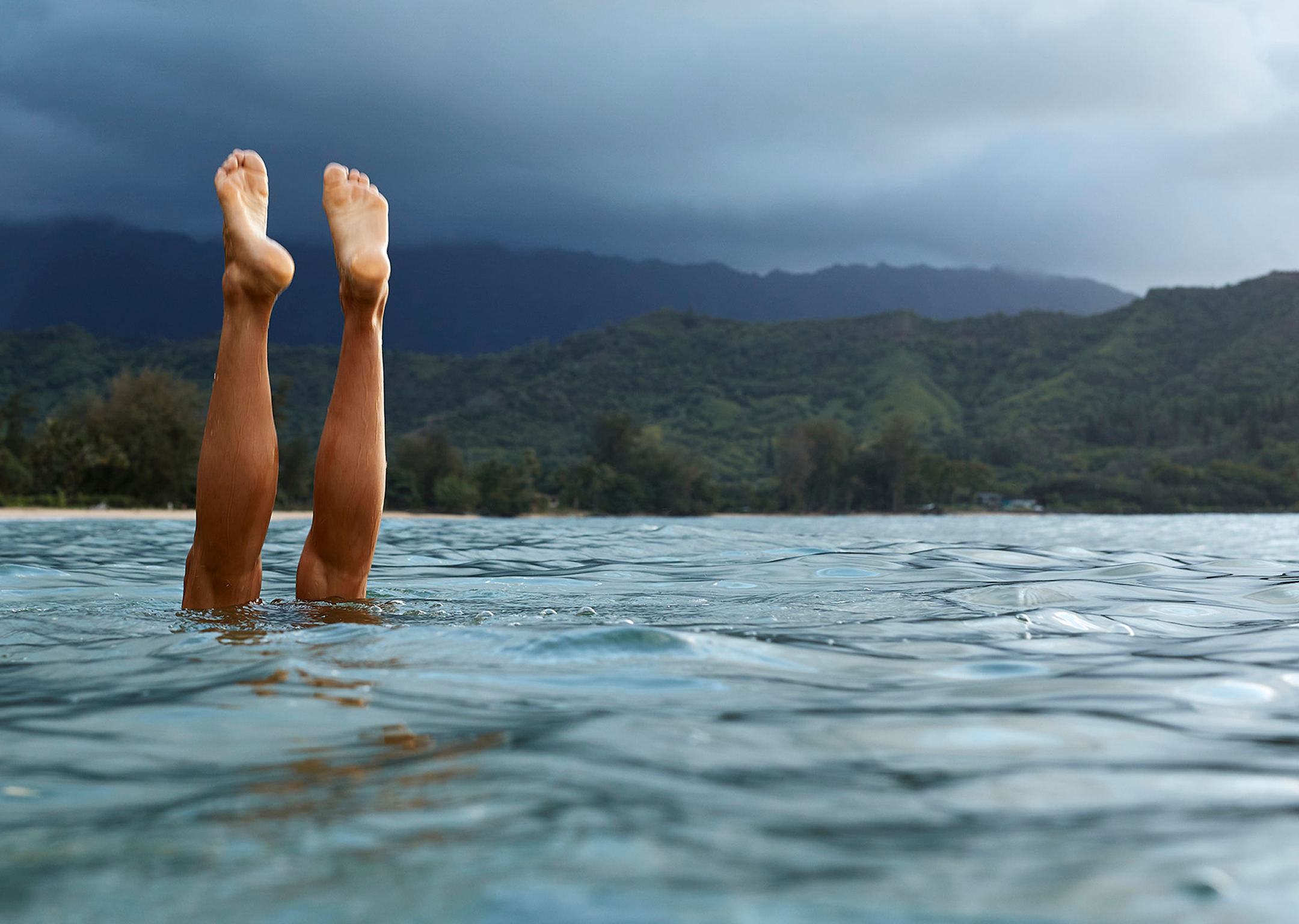 Person's legs sticking straight up out of the ocean water with a mountainous shoreline and cloudy sky in the background.