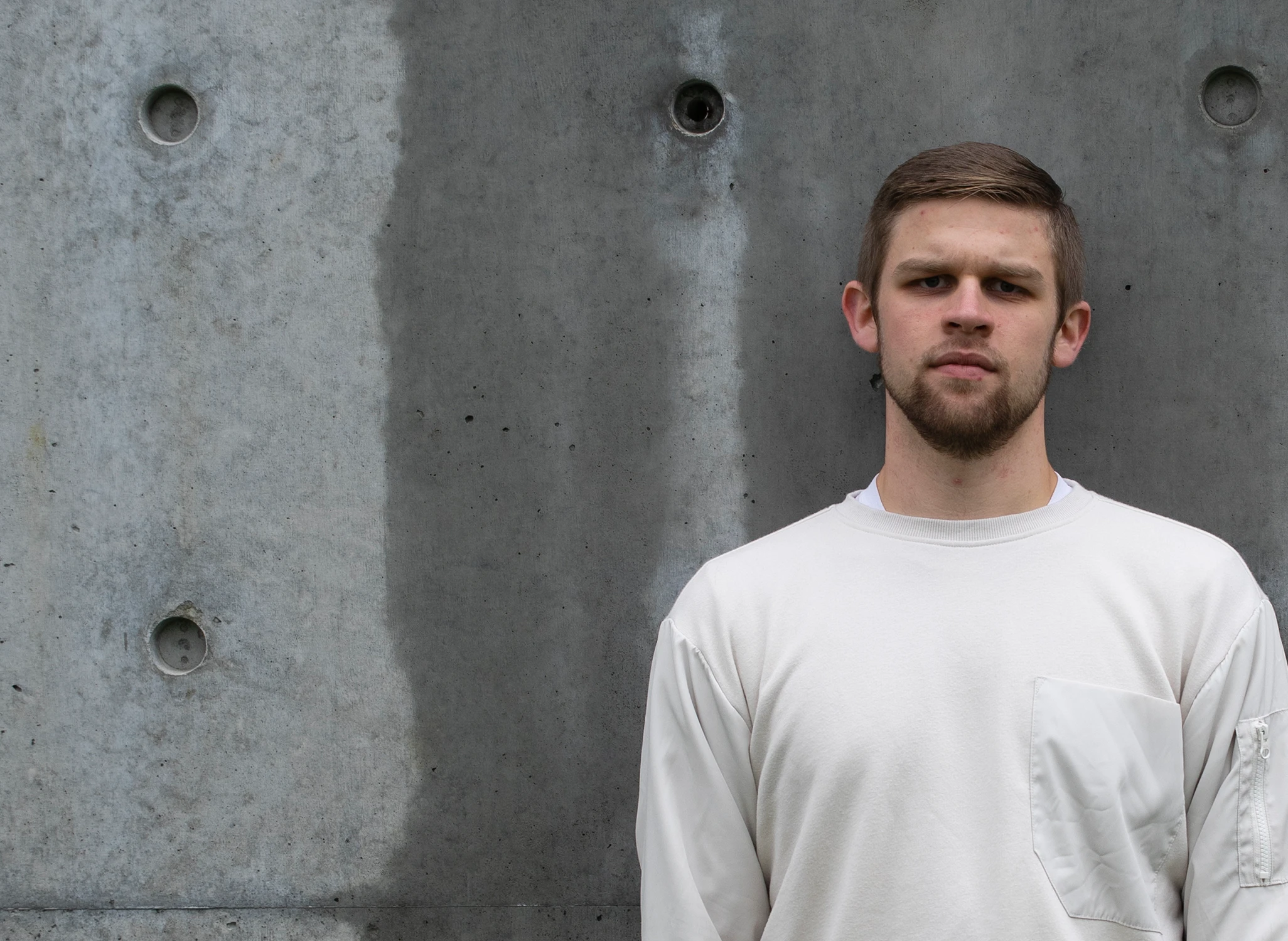 Stark image of a young man with short hair and a beard wearing a white sweatshirt standing with a serious expression against a textured concrete wall.