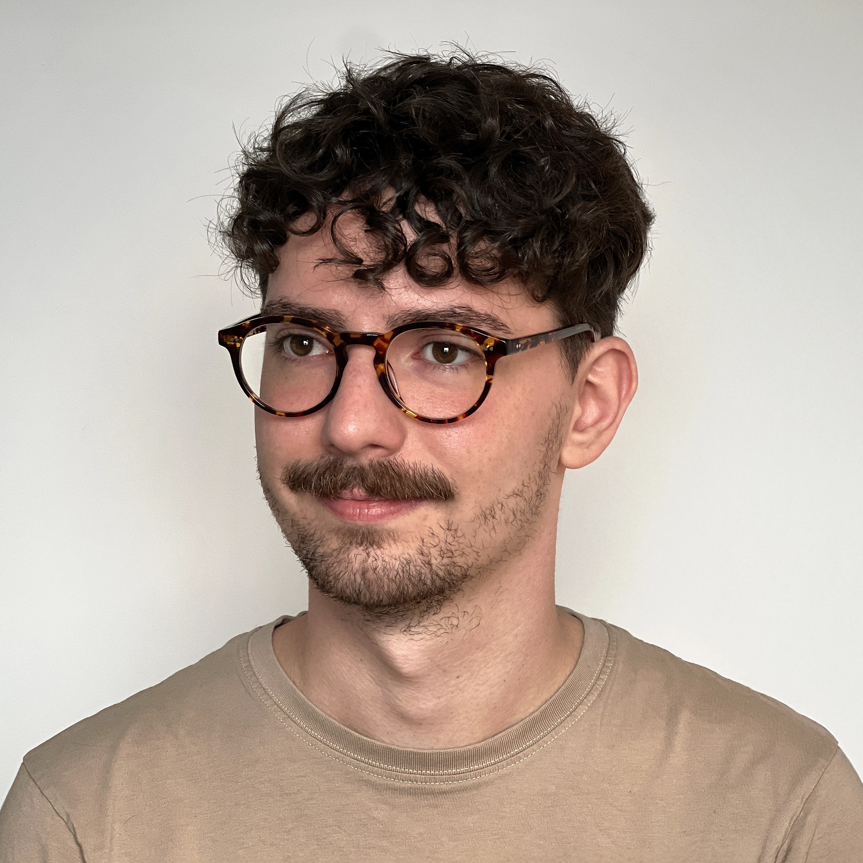 Headshot image of Pat Bullock; a man with curly dark hair, mustache, and tortoiseshell glasses wearing a beige t-shirt against a neutral off-white background.