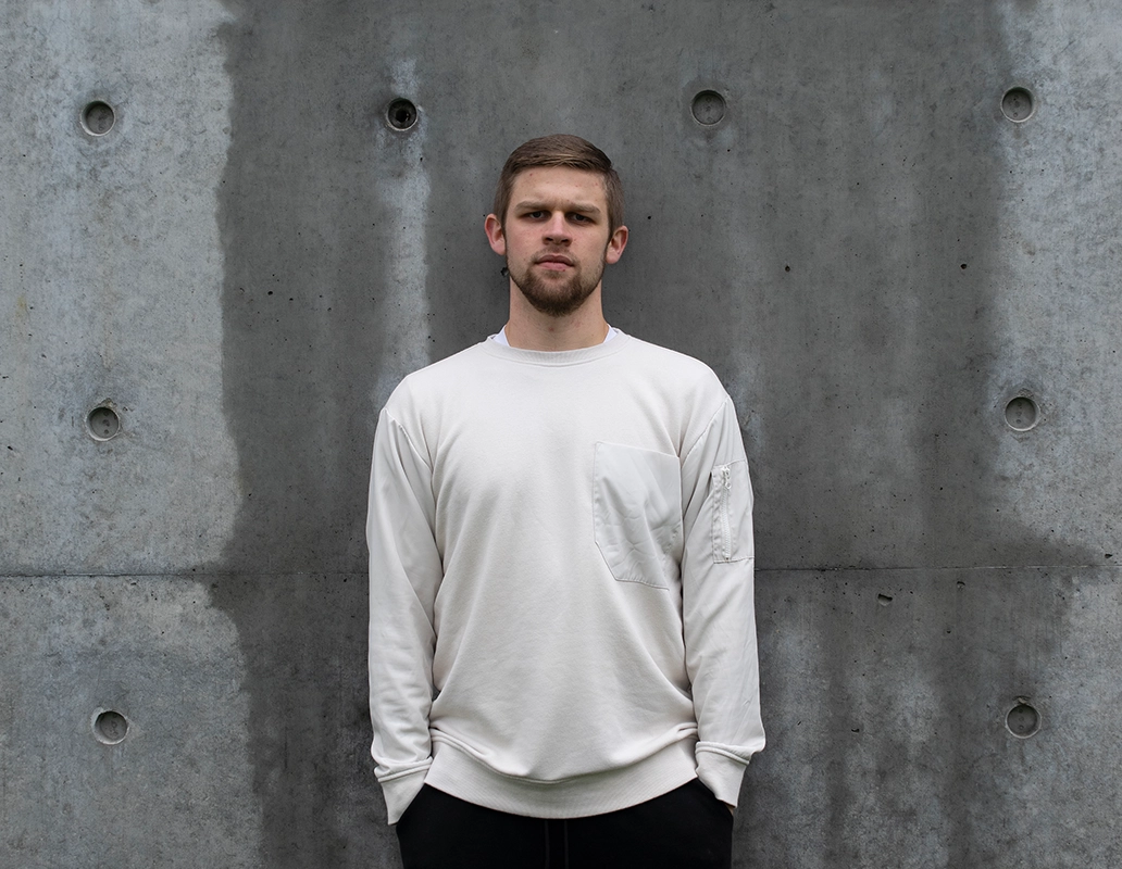Stark image of a young man with short hair and a beard wearing a white sweatshirt standing with a serious expression against a textured concrete wall.