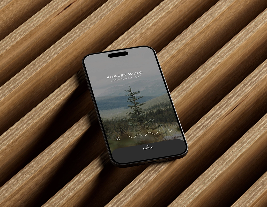 Smartphone displaying an app screen titled 'Forest Wind Johnsbach, AUT' with a pine tree and green mountain background, lying on a modern wooden slatted surface.