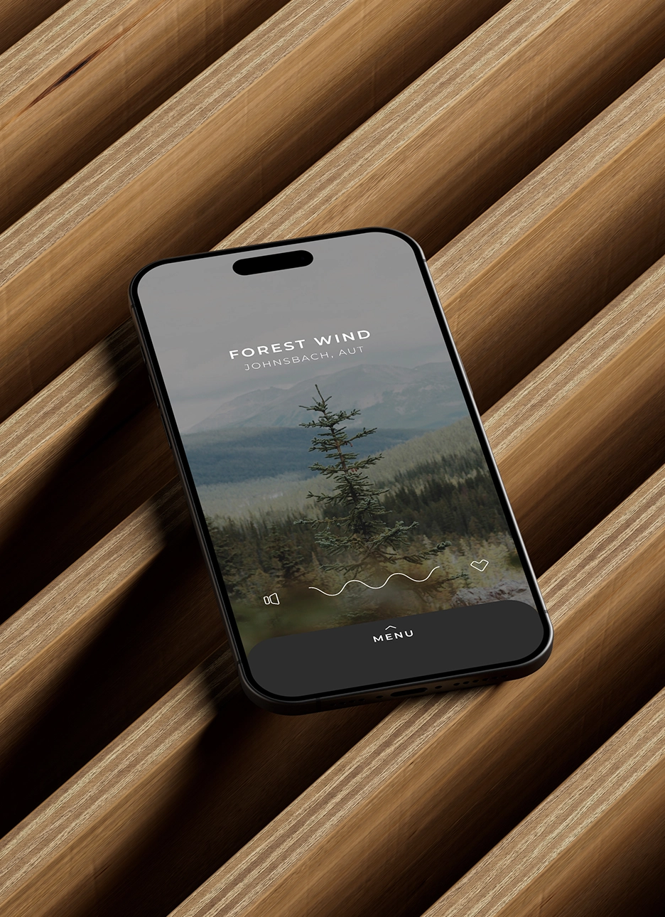 Smartphone displaying an app screen titled 'Forest Wind Johnsbach, AUT' with a pine tree and green mountain background, lying on a modern wooden slatted surface.