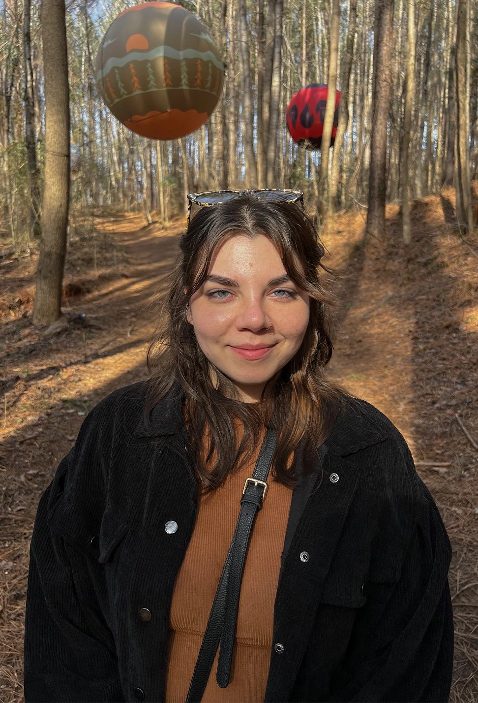 Portrait photo of a young woman with brown hair and blue eyes standing on a barren but warm forest path with two decorative balloons floating behind her.