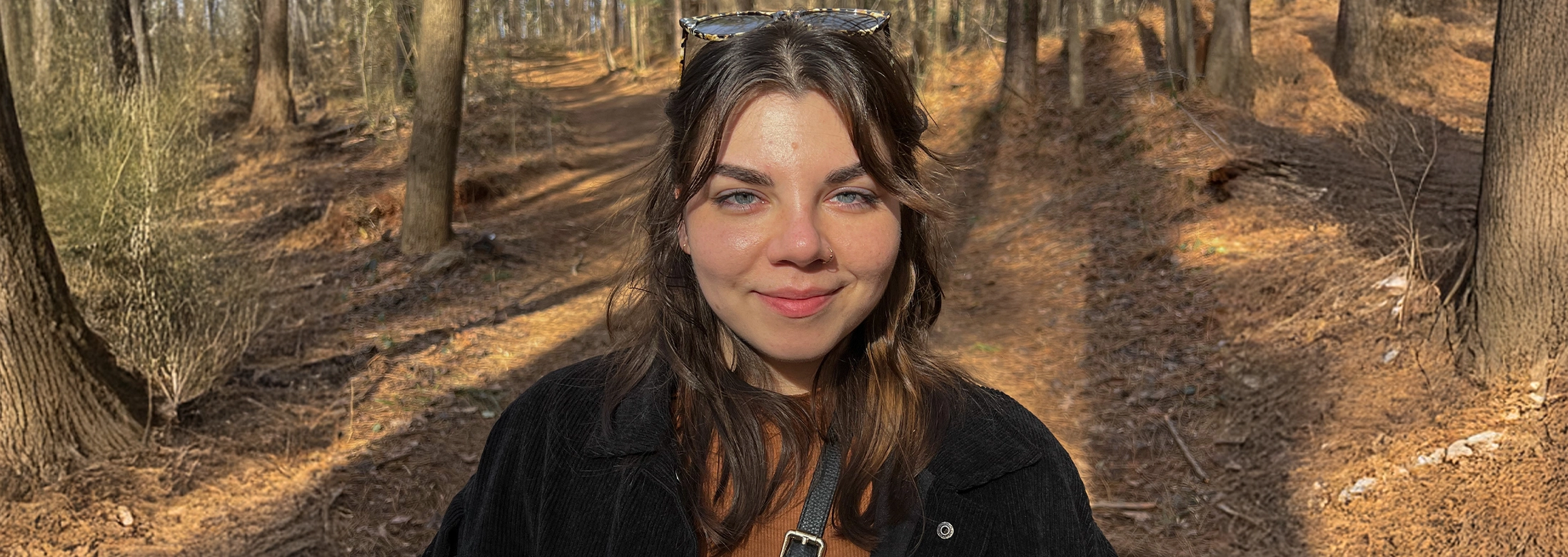 Portrait photo of a young woman with brown hair and blue eyes standing on a barren but warm forest path.