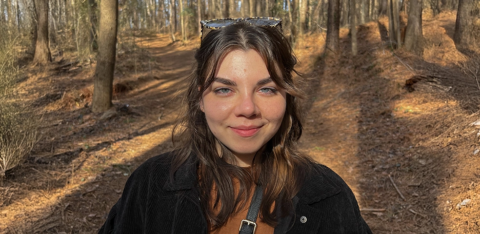 Portrait photo of a young woman with brown hair and blue eyes standing on a barren but warm forest path.