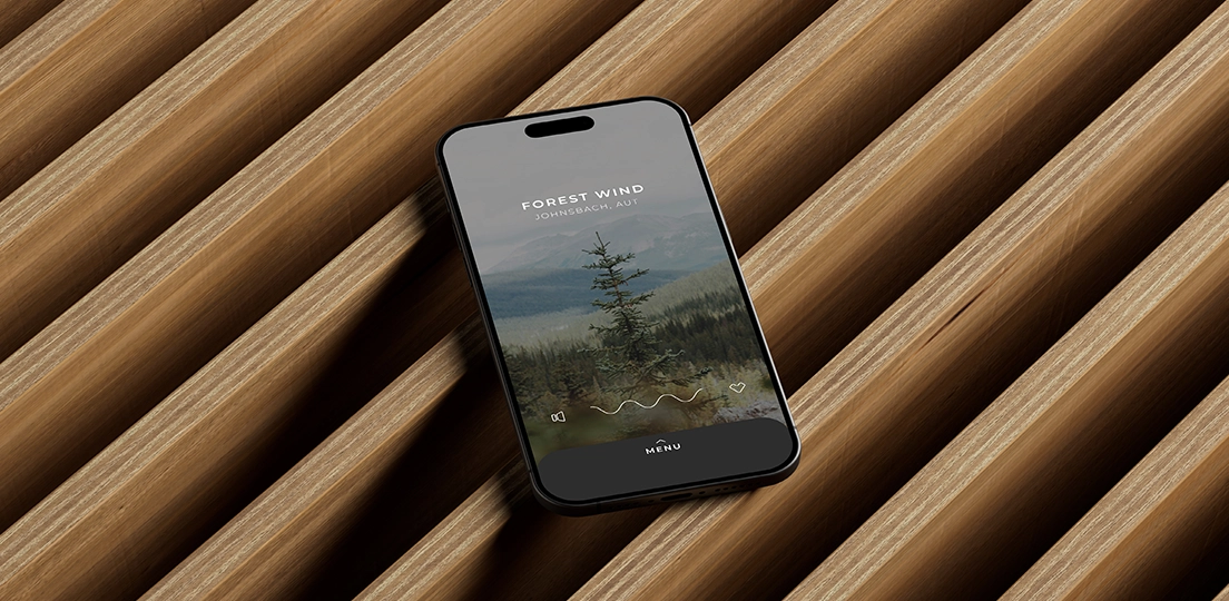 Smartphone displaying an app screen titled 'Forest Wind Johnsbach, AUT' with a pine tree and green mountain background, lying on a modern wooden slatted surface.
